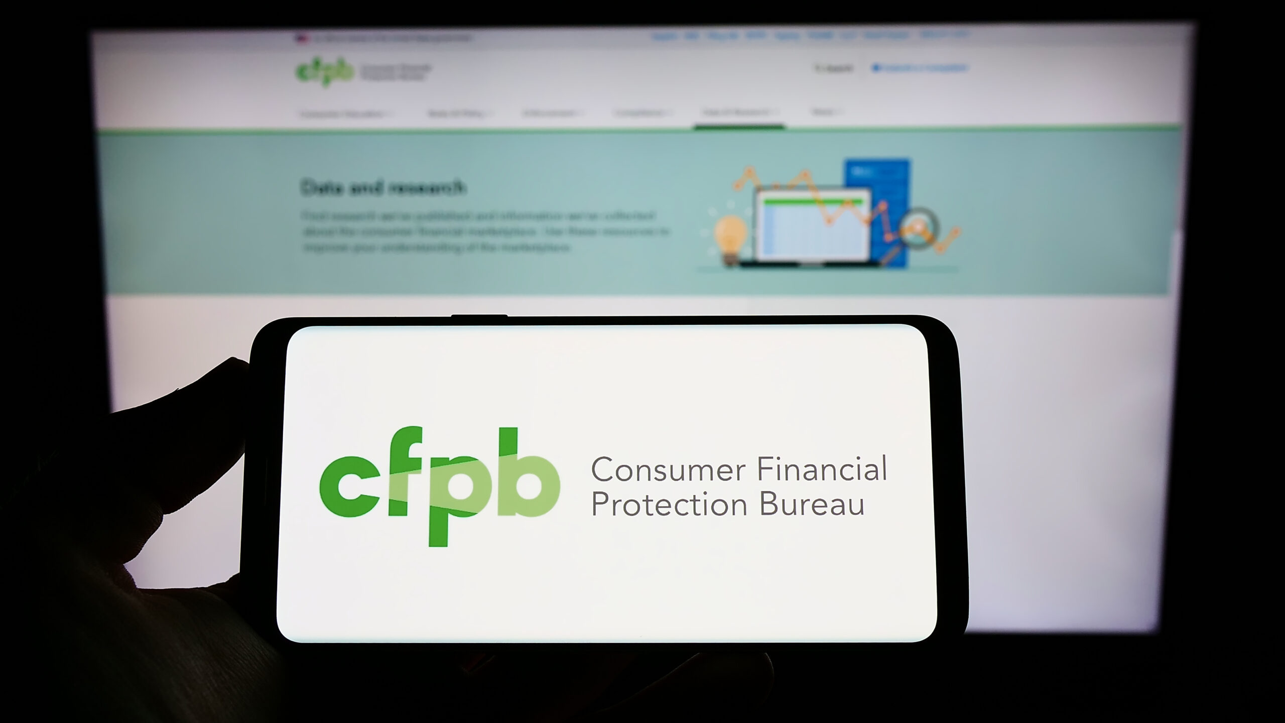 Person holding smartphone with logo of US Consumer Financial Protection Bureau (CFPB) on screen in front of website. Focus on phone display.